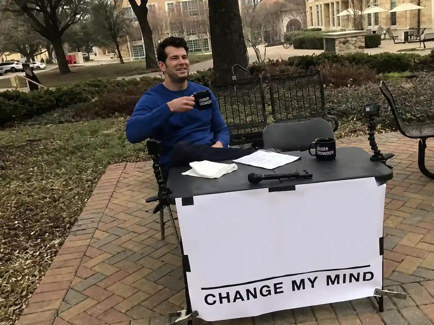 A man sitting at a table with a coffee mug and a large sign that reads Change My Mind.