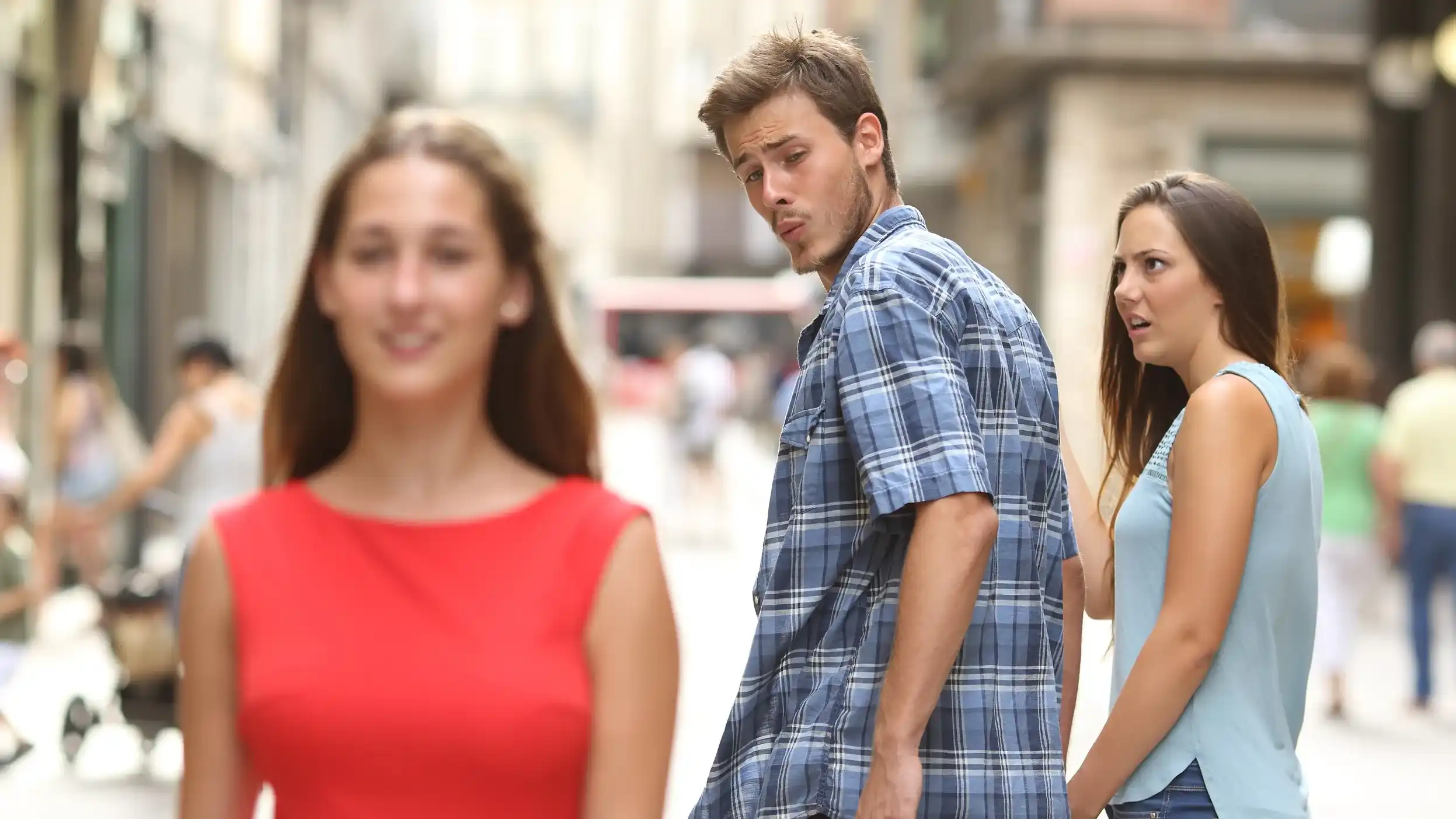 A man walks with his girlfriend but turns around to look at another woman in a red dress as his girlfriend looks at him in shock.